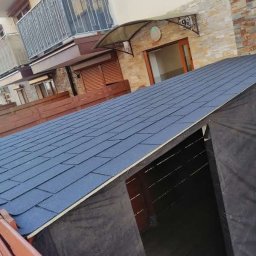 Stolkra - Wznoszona altana z ciemnym gontem bitumicznym na dachu, widoczna na tle budynku mieszkalnego z balkonami i drewnianym ogrodzeniem, w trakcie budowy.