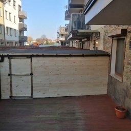 Stolkra - Drewniana altana z drzwiami na tarasie z desek w otoczeniu budynków mieszkalnych. Widok na miasto w tle.