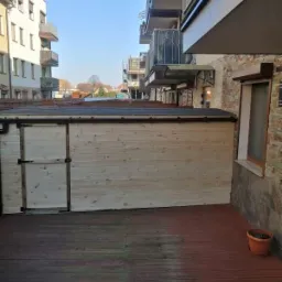 Drewniana altana z drzwiami na tarasie z desek w otoczeniu budynków mieszkalnych. Widok na miasto w tle.