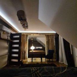 Stolkra - Drewniana aranżacja poddasza z oświetleniem LED, widoczna lampka na biurku i ciemne zasłony. Styl rustykalny z nowoczesnymi akcentami w wykończeniu wnętrza.