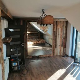 Stolkra - Drewniane wykończenie wnętrza z lampą w stylu rustykalnym. Widoczna podłoga z paneli i ściany obłożone drewnem. W tle schody i okno.