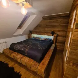 Rustykalna sypialnia na poddaszu: drewniane panele ścienne, masywne łóżko z bali, oświetlenie żarówkami Edisona. Widoczne skosy i okno dachowe.