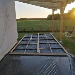 Stolkra - Budowa tarasu: drewniany szkielet z wypełnieniem żwirowym na folii, konstrukcja z belek pod zadaszeniem. W tle zielone pole w promieniach zachodzącego słońca.