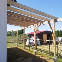 Stolkra - Konstrukcja z surowego drewna altany w trakcie budowy, widok z boku. Słupy osadzone na betonowych podstawach, w tle zielona trawa i budynek z czerwonym dachem.