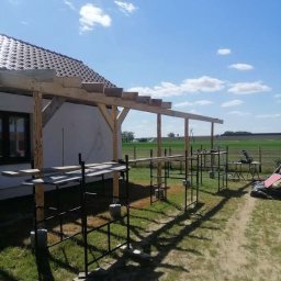 Stolkra - Drewniana konstrukcja zadaszenia tarasu w trakcie budowy przy domu jednorodzinnym, z widocznymi rusztowaniami i elementami roboczymi. Słoneczny dzień.