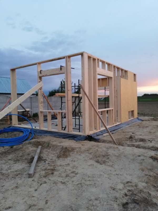 Wznoszenie drewnianej konstrukcji szkieletowej domu na fundamencie, widoczne elementy rusztowania i niebieski przewód. Budowa w toku, tło: zachodzące słońce.