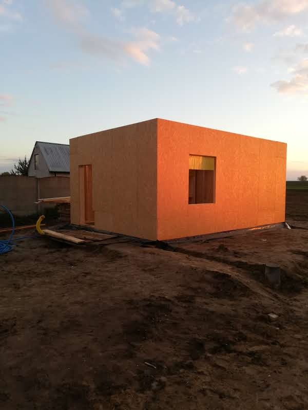 Wczesny etap budowy domu szkieletowego z widoczną konstrukcją ścian z płyty OSB, otworem okiennym i drzwiowym na tle zachodzącego słońca.