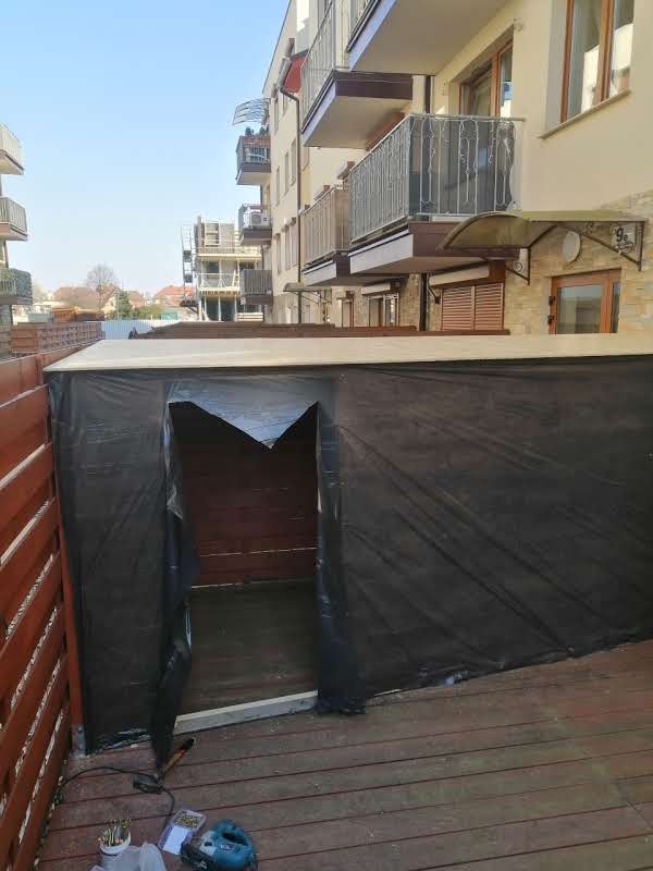 Montaż zadaszenia na tarasie z desek, zabezpieczonego czarną folią. W tle budynek mieszkalny z balkonami. Widoczne narzędzia pracy.