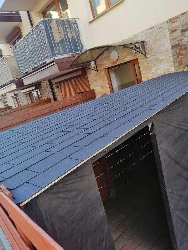 Wznoszona altana z ciemnym gontem bitumicznym na dachu, widoczna na tle budynku mieszkalnego z balkonami i drewnianym ogrodzeniem, w trakcie budowy.