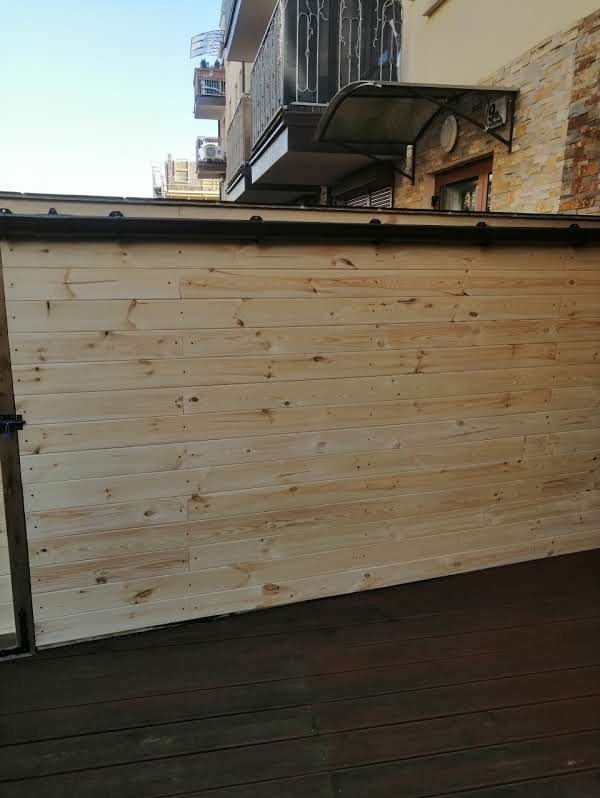 Drewniana zabudowa tarasu z jasnych desek na tle budynku z balkonami i zadaszeniem. Ciemna podłoga tarasu. Widok z perspektywy osoby stojącej na tarasie.