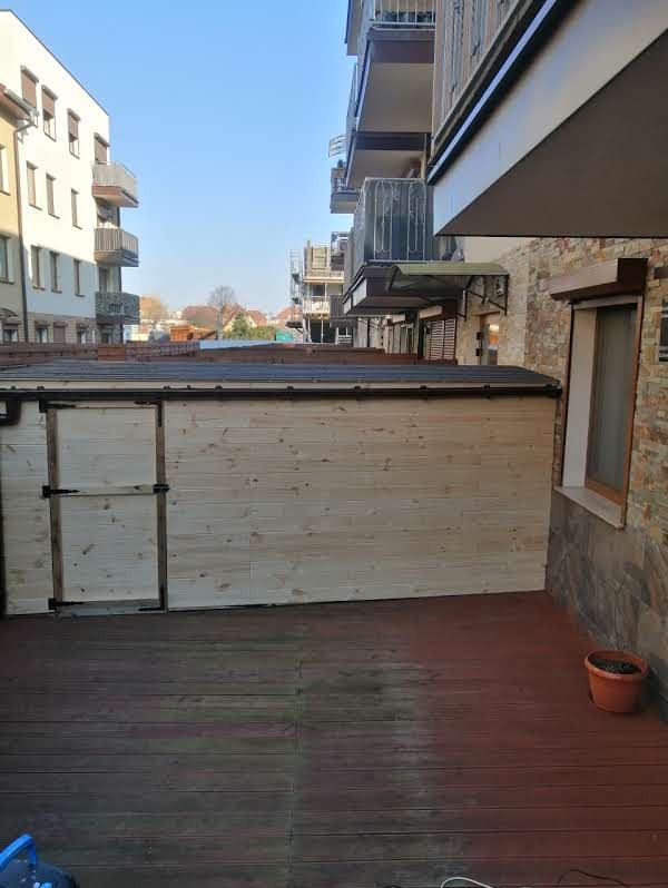 Drewniana altana z drzwiami na tarasie z desek w otoczeniu budynków mieszkalnych. Widok na miasto w tle.