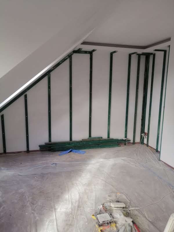 Szkieletowa konstrukcja ścian poddasza z zielonymi profilami, gotowa do obłożenia płytami. Podłoga zabezpieczona folią malarską, widoczne narzędzia.