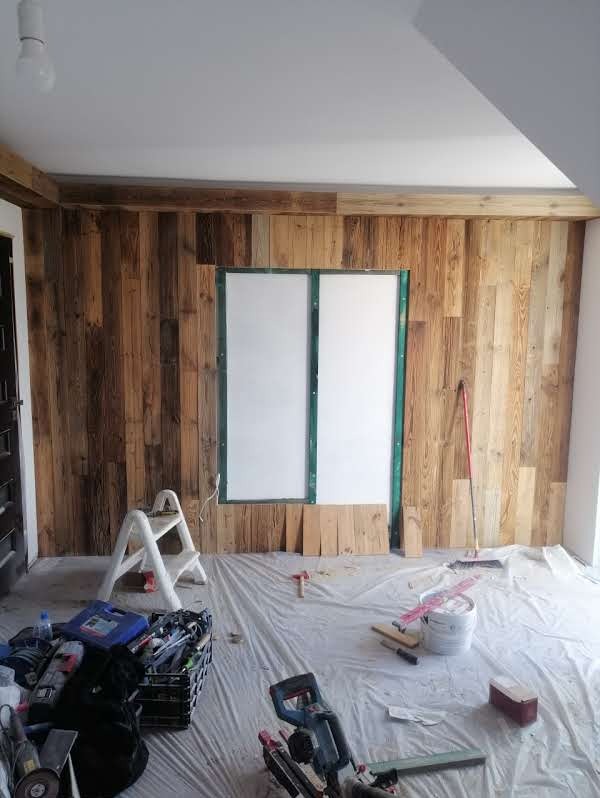 Montaż boazerii drewnianej w pomieszczeniu: deski przy oknie, drabina, narzędzia na folii ochronnej. Widoczne etapy prac wykończeniowych. Ciepłe odcienie drewna.