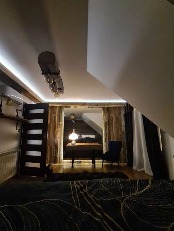Drewniana aranżacja poddasza z oświetleniem LED, widoczna lampka na biurku i ciemne zasłony. Styl rustykalny z nowoczesnymi akcentami w wykończeniu wnętrza.