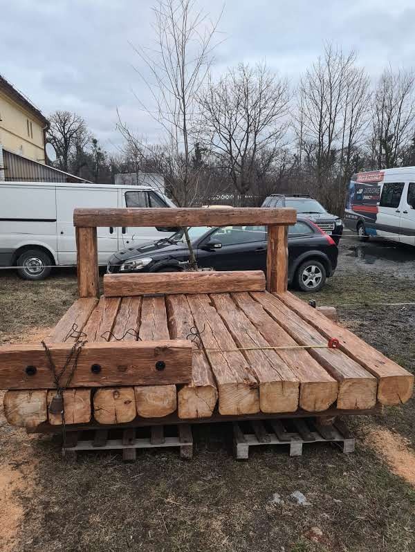 Surowa konstrukcja altany z bali, widoczne elementy mocujące i wstępny montaż na palecie, otoczenie z pojazdami i budynkami w tle.