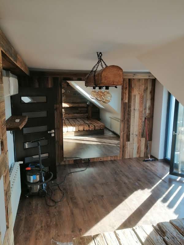 Drewniane wykończenie wnętrza z lampą w stylu rustykalnym. Widoczna podłoga z paneli i ściany obłożone drewnem. W tle schody i okno.