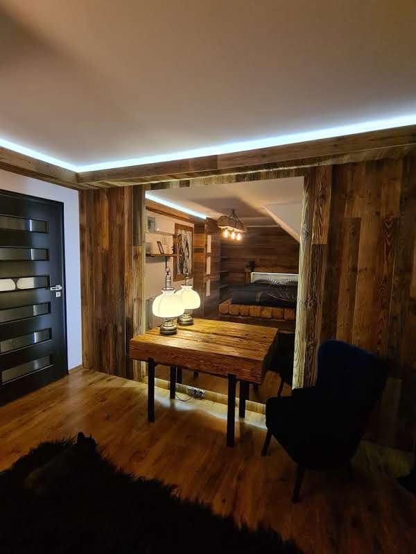 Drewniane wnętrze z podświetlanym sufitem, stolikiem i widokiem na sypialnię. Ciepłe oświetlenie i naturalne materiały tworzą przytulną atmosferę.