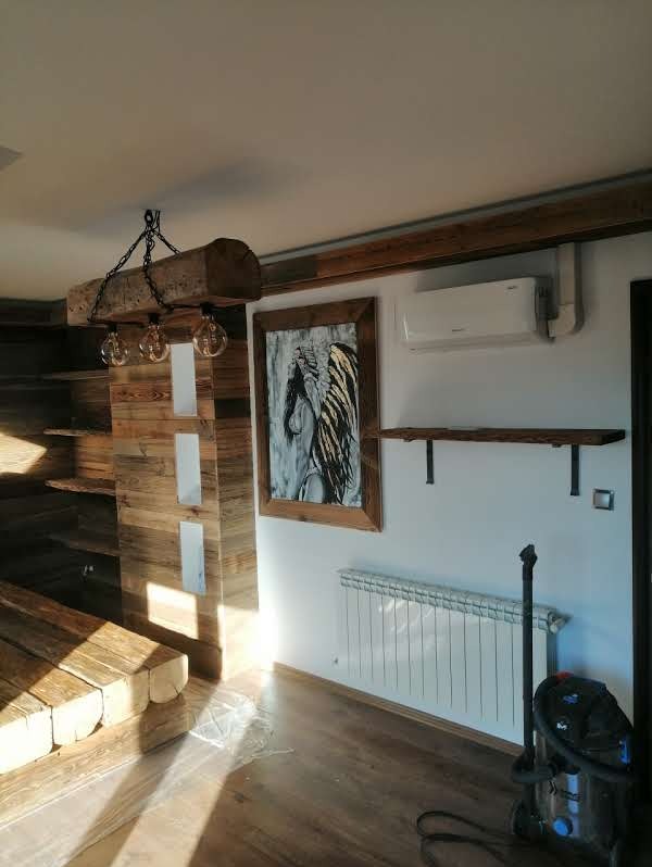 Drewniane schody z surowych bali i lampa z belki w rustykalnym wnętrzu. Na ścianie obraz w ramie i grzejnik. Widoczny odkurzacz.