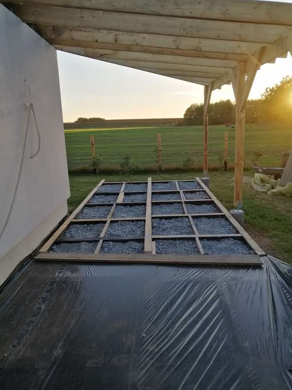 Budowa tarasu: drewniany szkielet z wypełnieniem żwirowym na folii, konstrukcja z belek pod zadaszeniem. W tle zielone pole w promieniach zachodzącego słońca.