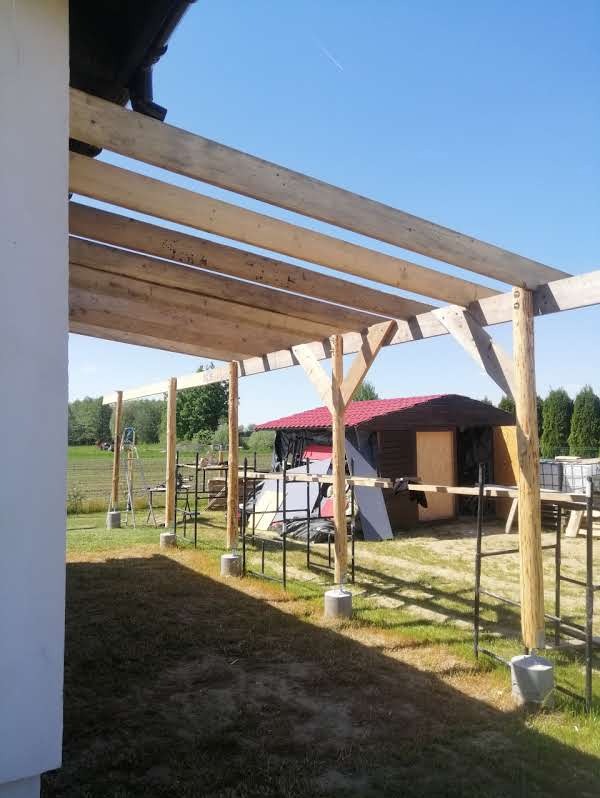 Konstrukcja z surowego drewna altany w trakcie budowy, widok z boku. Słupy osadzone na betonowych podstawach, w tle zielona trawa i budynek z czerwonym dachem.