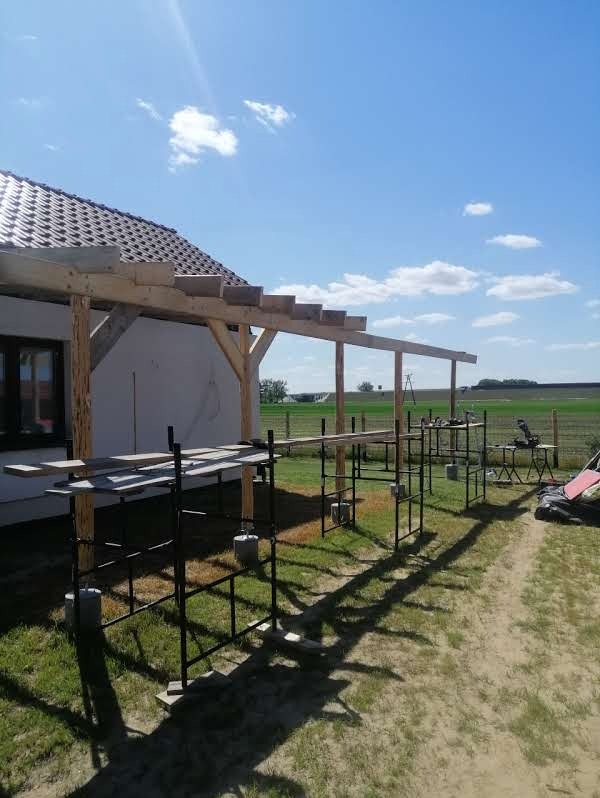 Drewniana konstrukcja zadaszenia tarasu w trakcie budowy przy domu jednorodzinnym, z widocznymi rusztowaniami i elementami roboczymi. Słoneczny dzień.