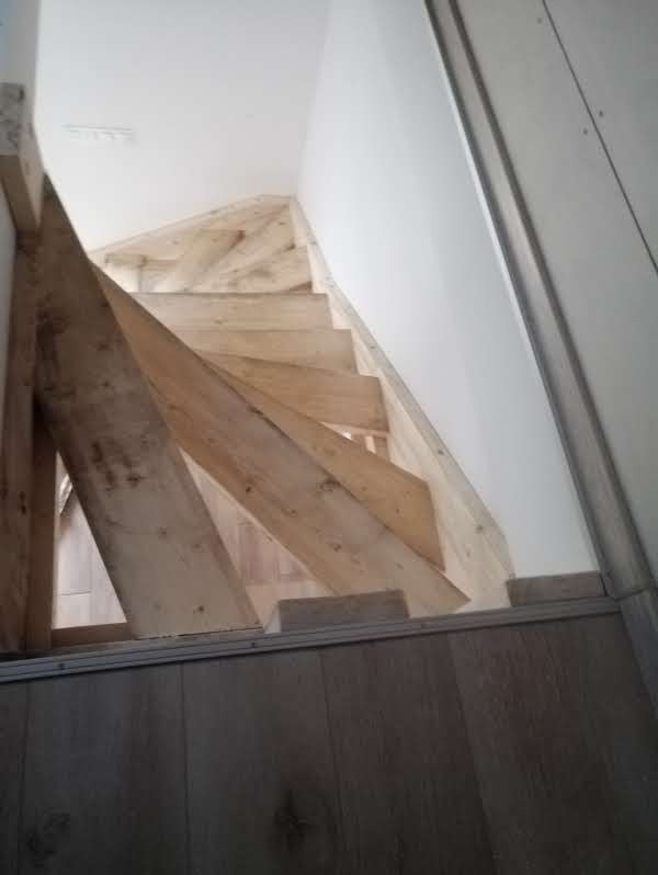 Surowe drewniane schody w trakcie montażu, widoczne elementy konstrukcyjne. Ściana wykończona płytą, podłoga z paneli. Ujęcie z dołu, podkreślające etap budowy.