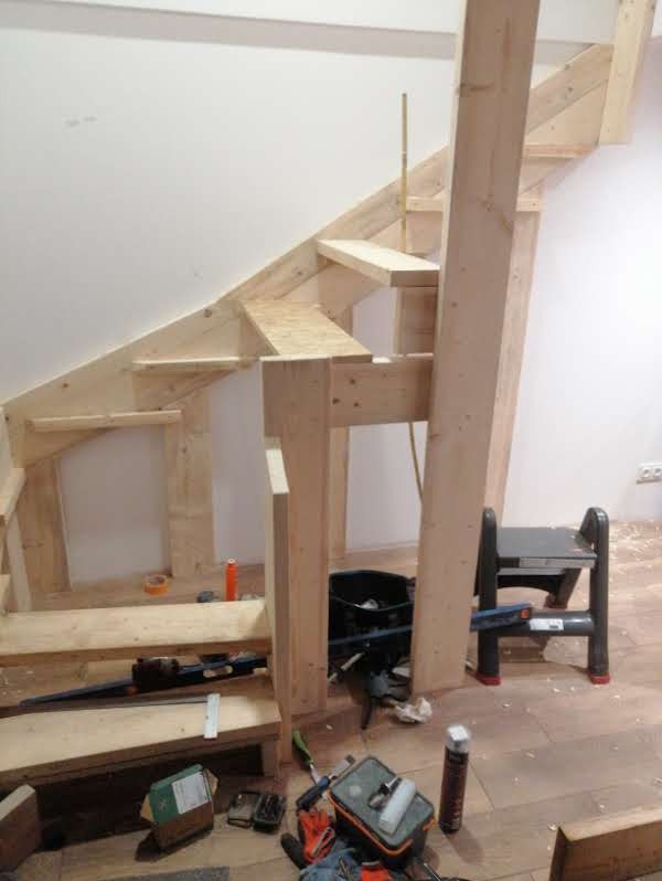 Surowe drewniane schody w trakcie budowy, widoczne narzędzia stolarskie i drabinka. Elementy konstrukcyjne w jasnym drewnie na tle białej ściany.