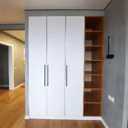 Biała szafa z trzema drzwiami i długimi, czarnymi uchwytami, z otwartą, drewnianą półką po prawej stronie, na tle szarej ściany i drewnianej podłogi.