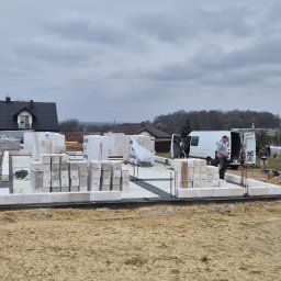 MAR-BUD Marcin Świerkosz - Budowa domu: wzniesione fundamenty z bloczków, przygotowane do dalszych prac murarskich; widoczny pracownik przy samochodzie dostawczym na tle krajobrazu.