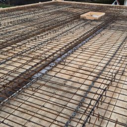 MAR-BUD Marcin Świerkosz - Zbrojenie stropu na drewnianym szalunku z widocznymi dystansami z tworzywa sztucznego, przygotowane do zalania betonem, na tle zabudowy jednorodzinnej i pochmurnego nieba.