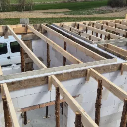 Wznoszenie ścian domu z bloczków, widoczne drewniane podpory stropu z nieobrobionych pni drzew, w tle krajobraz pól i biały samochód dostawczy.