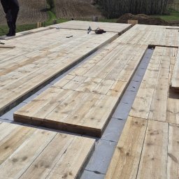 MAR-BUD Marcin Świerkosz - Wczesny etap budowy domu: widok na fundamenty z szarych bloczków betonowych, na których ułożono drewniane deski pod podłogę, z narzędziami budowlanymi w tle i fragmentem sylwetki pracownika.