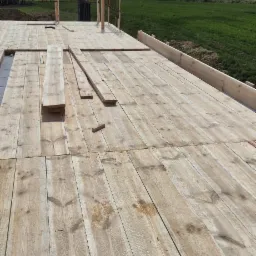 Drewniana konstrukcja szalunku fundamentów domu jednorodzinnego z widokiem na zielone pola i zabudowania w tle, w słoneczny dzień.