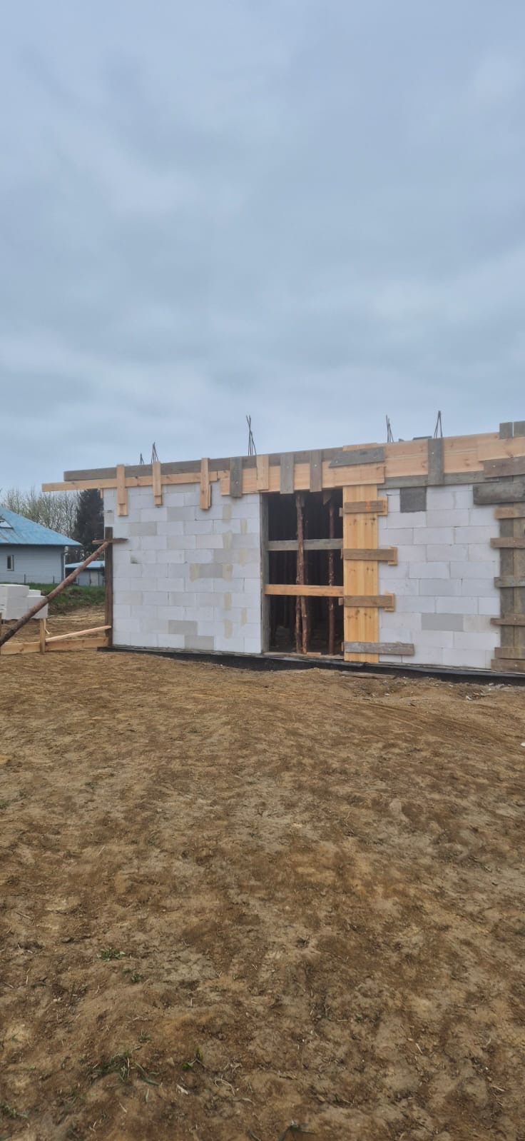 Wznoszenie ścian parterowego domu z bloczków betonowych, widoczne drewniane szalunki i podpory, w tle pochmurne niebo i fragment budynku.