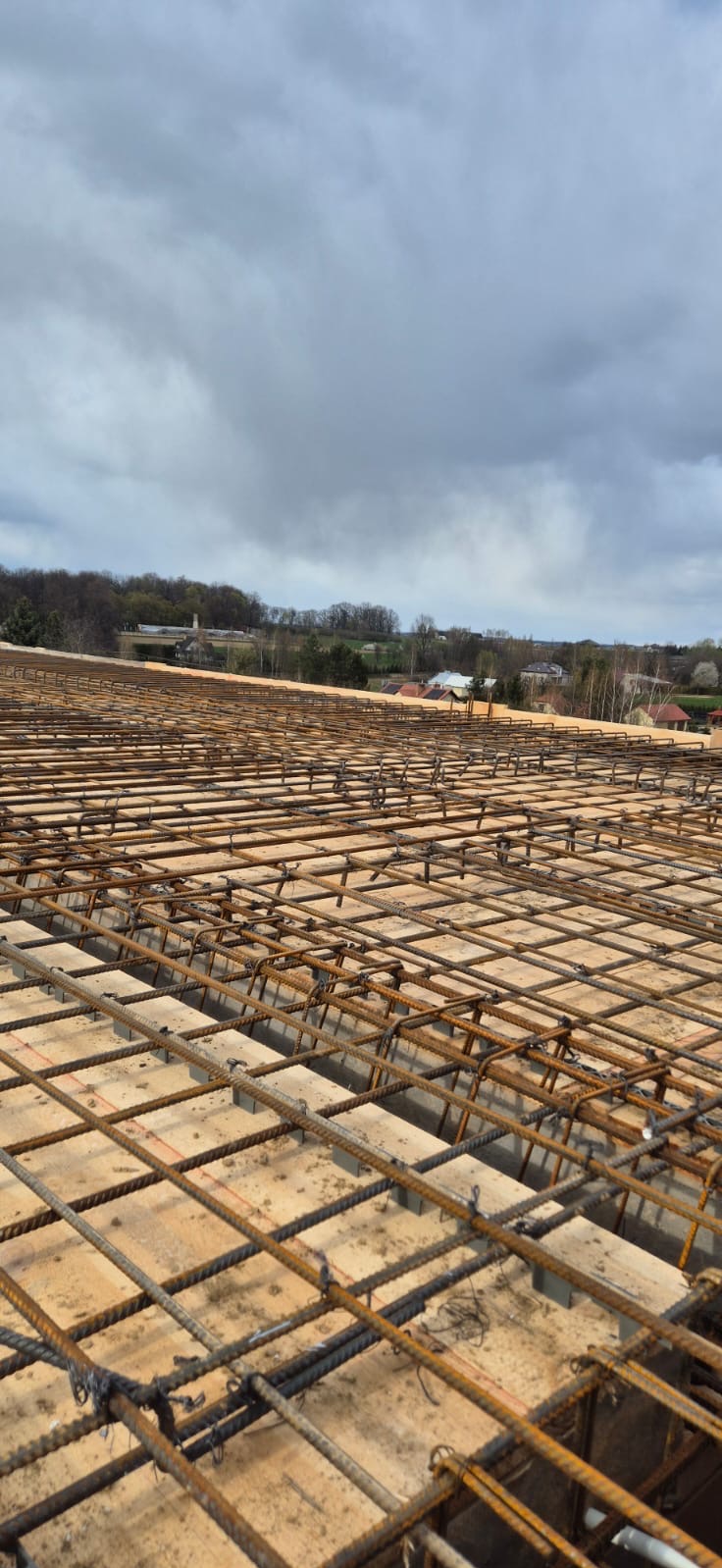 Zbrojenie płyty fundamentowej z widocznymi dystansami i elementami wiązań, w tle krajobraz z zabudowaniami pod pochmurnym niebem.
