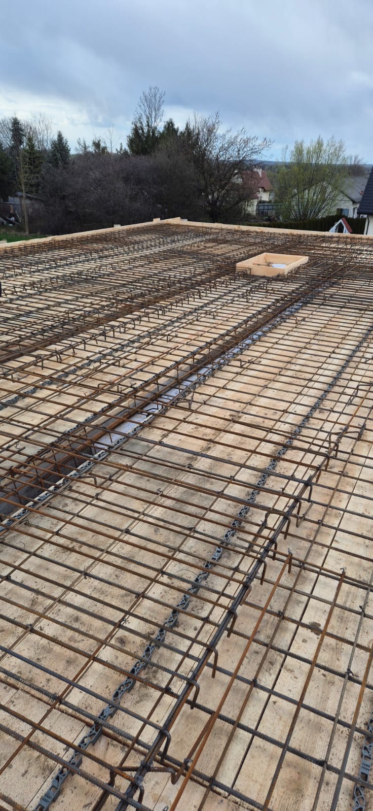 Zbrojenie stropu na drewnianym szalunku z widocznymi dystansami z tworzywa sztucznego, przygotowane do zalania betonem, na tle zabudowy jednorodzinnej i pochmurnego nieba.