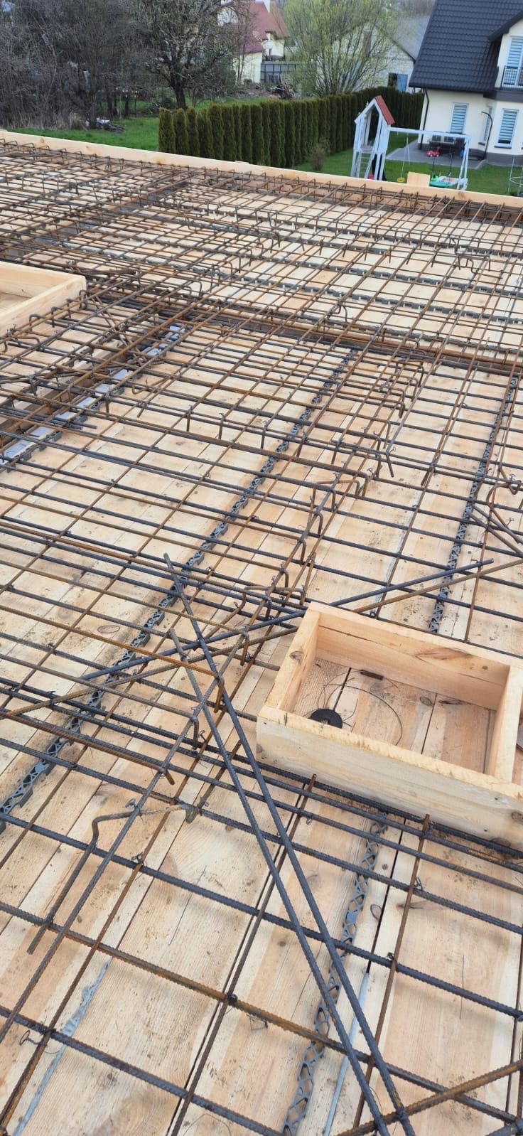 Siatka zbrojeniowa ułożona na deskowaniu stropu, widoczne drewniane szalunki i elementy dystansowe, w tle dom jednorodzinny i zieleń.