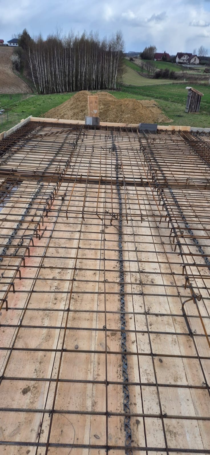 Zbrojenie płyty fundamentowej ułożone na deskowaniu, widok z góry na siatkę z prętów stalowych i dystanse z tworzywa sztucznego, w tle sterta ziemi i drzewa.