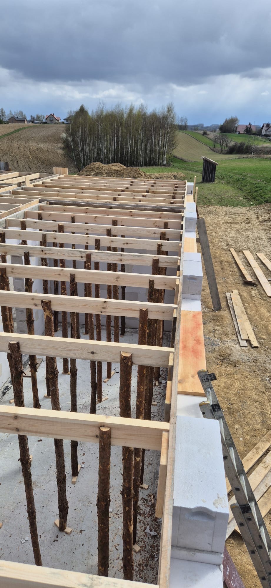 Wznoszenie ścian z bloczków betonowych, widoczne drewniane podpory i szalunki, przygotowanie do dalszych prac budowlanych.