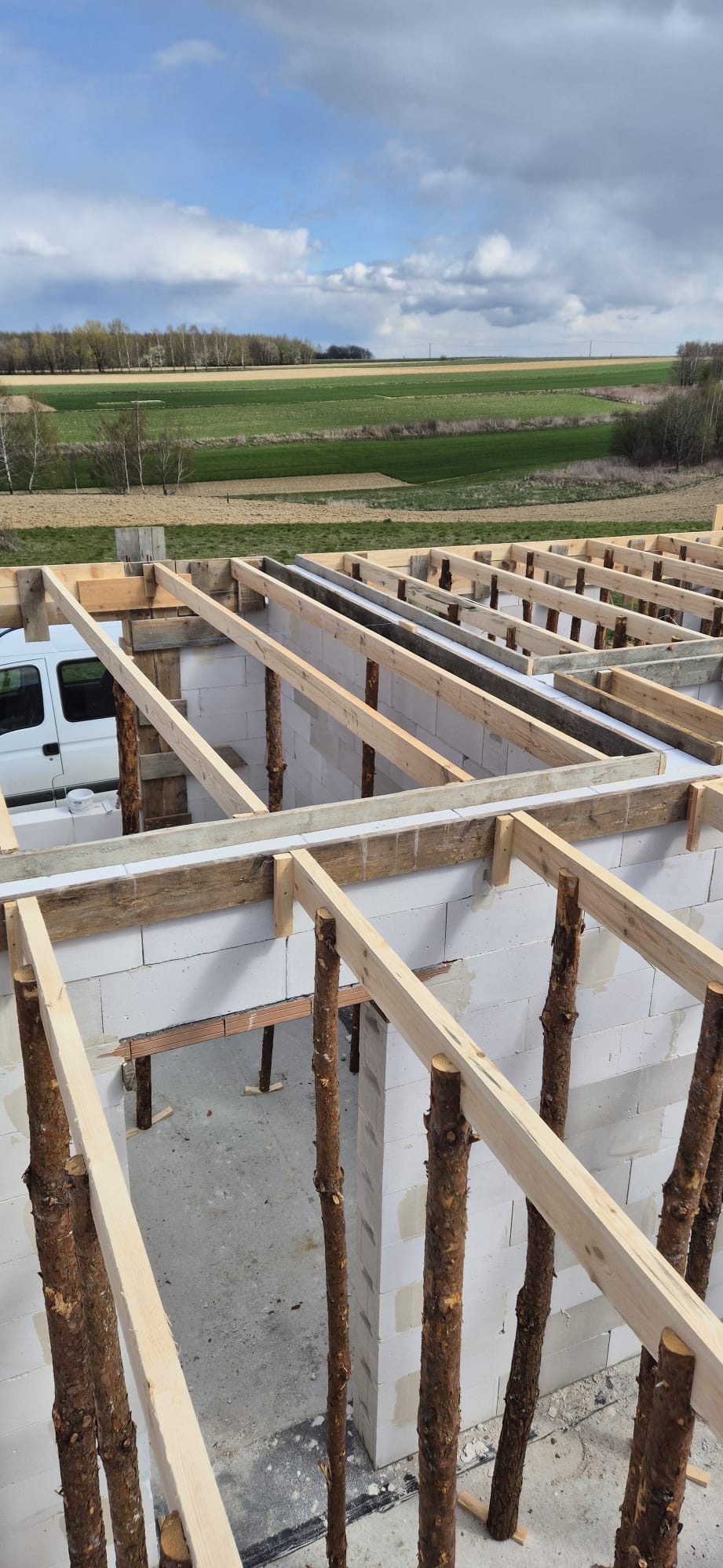 Wznoszenie ścian domu z bloczków, widoczne drewniane podpory stropu z nieobrobionych pni drzew, w tle krajobraz pól i biały samochód dostawczy.