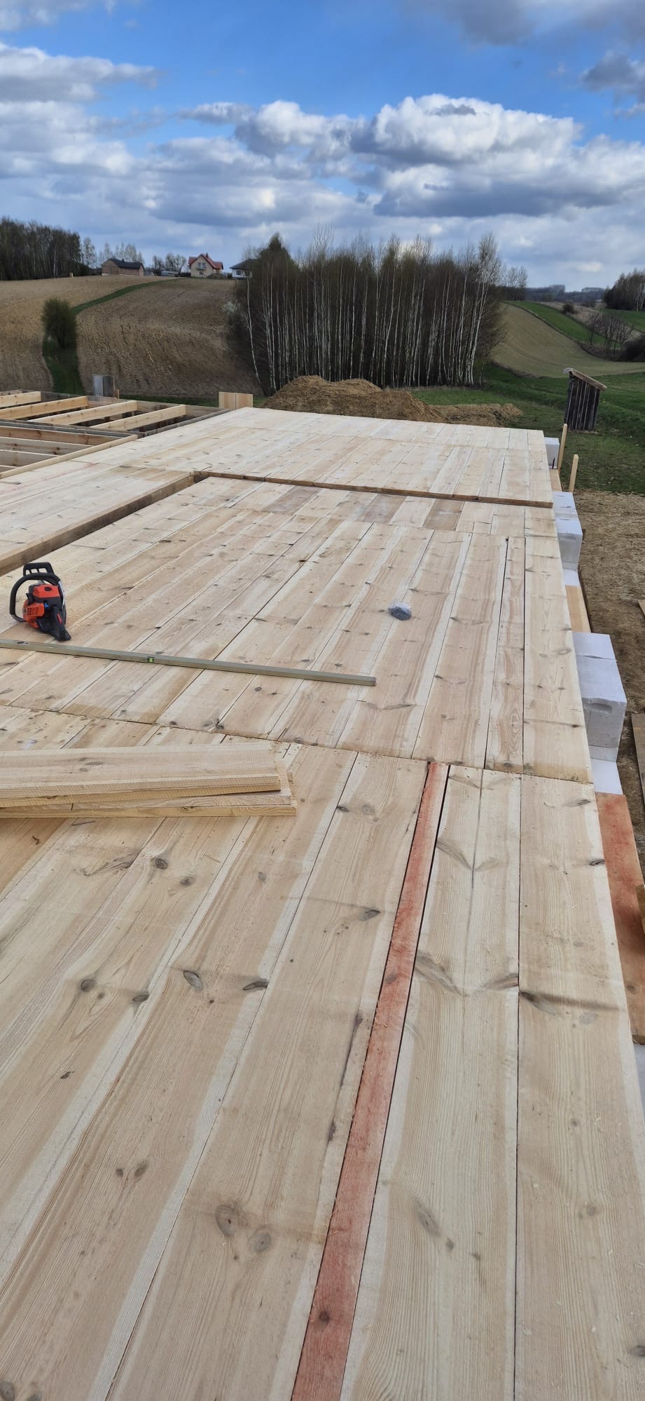 Wstępny etap budowy drewnianego stropu nad fundamentami, widoczna poziomica i piła spalinowa, w tle krajobraz wiejski.