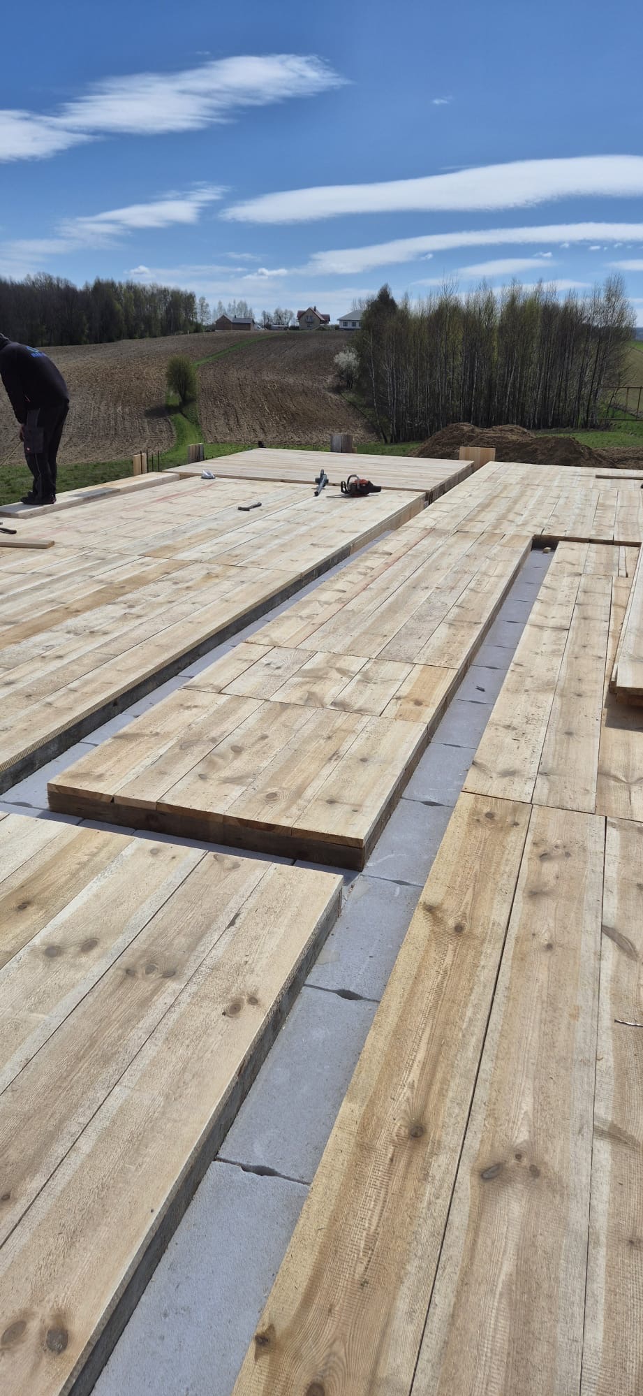 Wczesny etap budowy domu: widok na fundamenty z szarych bloczków betonowych, na których ułożono drewniane deski pod podłogę, z narzędziami budowlanymi w tle i fragmentem sylwetki pracownika.