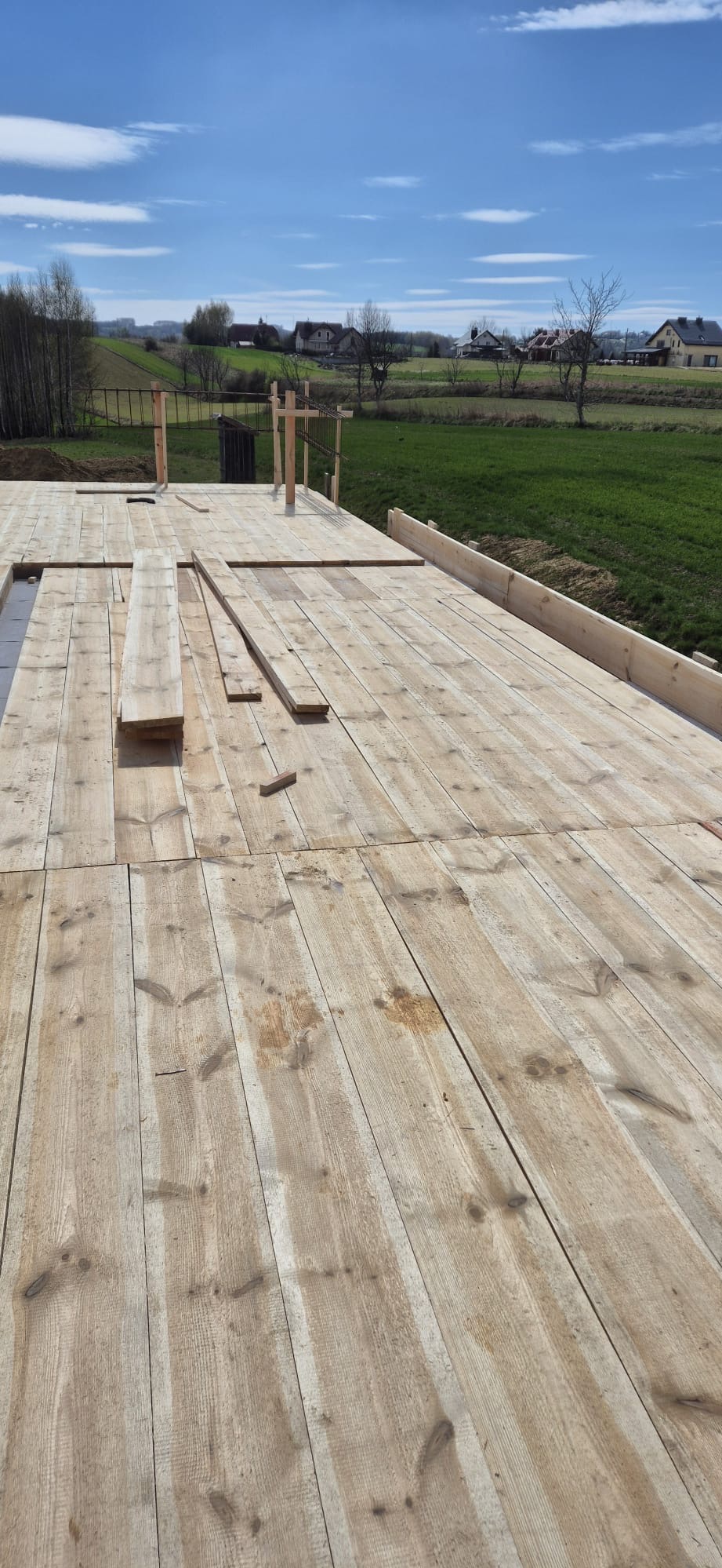 Drewniana konstrukcja szalunku fundamentów domu jednorodzinnego z widokiem na zielone pola i zabudowania w tle, w słoneczny dzień.