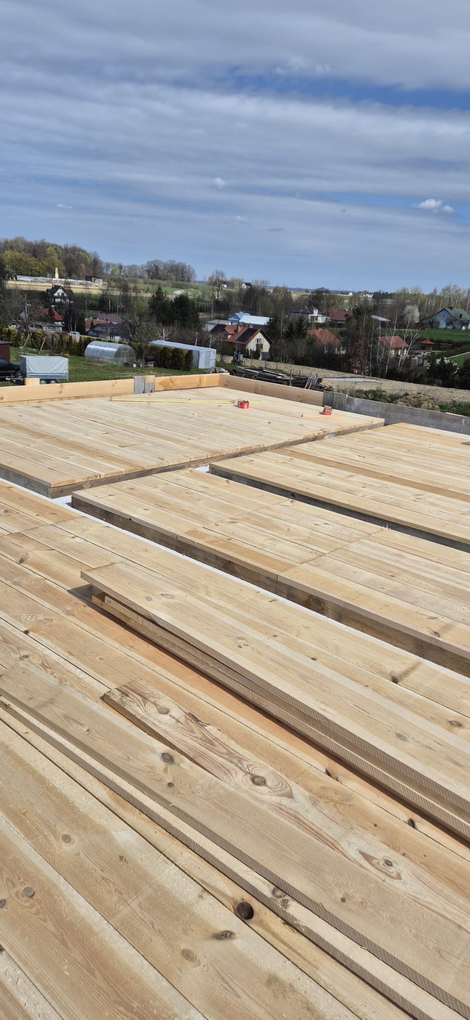 Wysoki kąt widzenia na drewniany strop nad fundamentami domu jednorodzinnego w trakcie budowy, widok na okoliczne domy i pola.