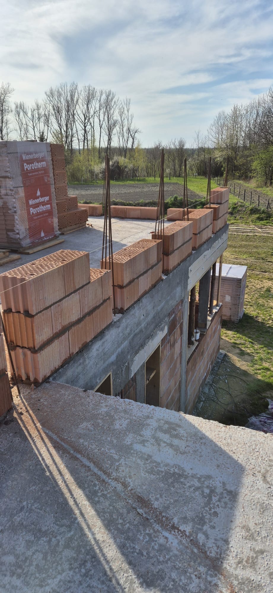 Wznoszenie murów domu z czerwonej cegły Porotherm, widoczne zbrojenie i paleta cegieł w folii, w tle drzewa i błękitne niebo.