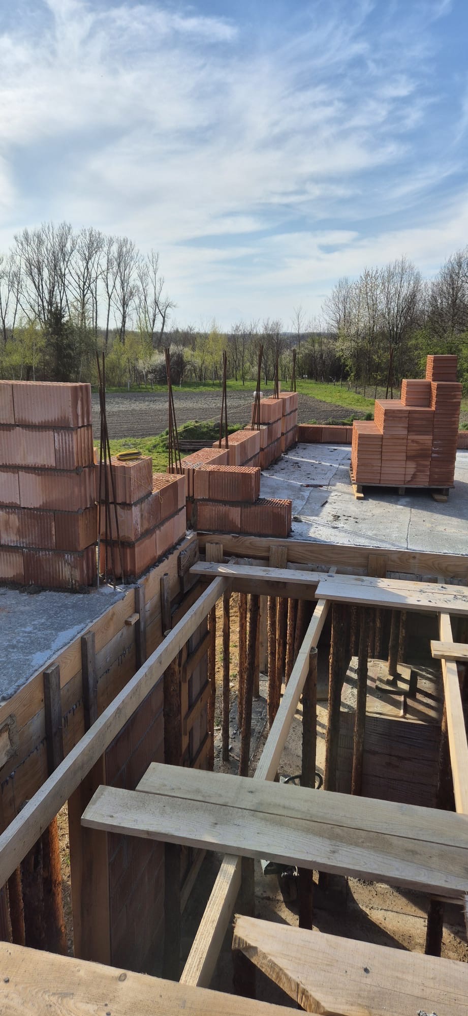 Wznoszenie ścian z bloczków ceramicznych Porotherm na fundamencie z drewnianym szalunkiem, widok na krajobraz polny w tle.