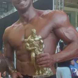 Uśmiechnięty kulturysta z brązową opalenizną, trzymający złotą statuetkę na tle sceny po zawodach Mens Physique. Widoczne mięśnie brzucha i ramion. Czerwone spodenki z czarnym pasem.