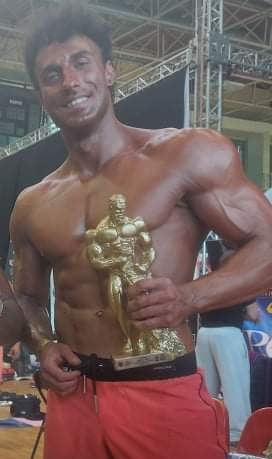 Uśmiechnięty kulturysta z brązową opalenizną, trzymający złotą statuetkę na tle sceny po zawodach Mens Physique. Widoczne mięśnie brzucha i ramion. Czerwone spodenki z czarnym pasem.