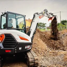 Biała mini koparka Bobcat E55 z operatorem w kabinie, podczas prac ziemnych na placu budowy; w tle widoczne domy jednorodzinne i ogrodzenie.