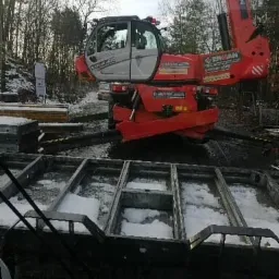 Czerwony dźwig teleskopowy marki Erman na placu budowy, przygotowujący podłoże pod fundamenty w zimowych warunkach. W tle widoczne drzewa i resztki śniegu.